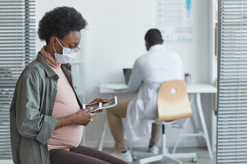 African pregnant woman in protective mask sitting and looking at ultrasound image in her hands during her visit at hospital