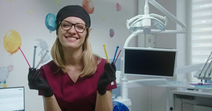 Young female doctor dentist orthodontist holding colored ligature rubber band for braces laughing risers in the dental office in a doctor's gown and hat
