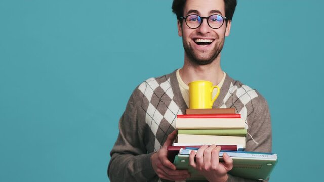 A happy man with glasses sitting down and standing up with a stack of books and a cup in a blue studio