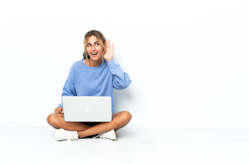 Young blonde Uruguayan girl with the laptop isolated on white background listening to something by putting hand on the ear