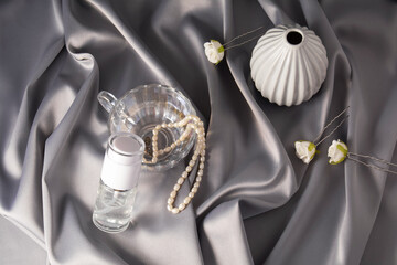 Luxury cosmetics concept. Still life on a shiny, delicate gray fabric. Beauty product with vase, flowers and glass form, pearl beads and hairpins with roses