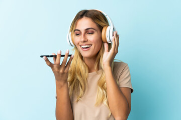 Naklejka premium Young blonde Uruguayan isolated on blue background listening music with a mobile and singing
