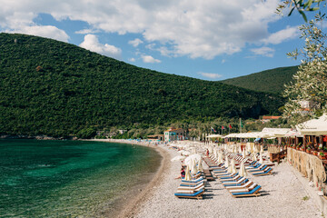 Fototapeta na wymiar View of the Zanjice beach on the Lustica peninsula in Montenegro against the backdrop of green mountains. Tourists rest on sun loungers