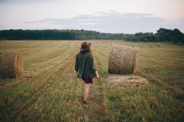 Naklejka na ściany i meble Beautiful stylish woman in hat walking at hay bales in summer evening field. Vacation in countryside