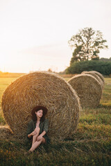 Naklejka na ściany i meble Beautiful woman in hat relaxing at haystacks enjoying evening sunset in summer field. Tranquility
