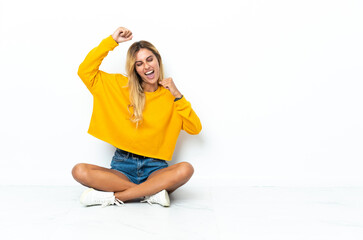 Fototapeta premium Young blonde Uruguayan woman sitting on the floor isolated on white background celebrating a victory