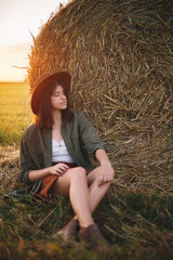 Naklejka na ściany i meble Beautiful woman in hat relaxing at haystacks enjoying evening sunset in summer field. Tranquility