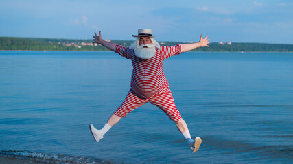 An elderly man in a striped retro swimsuit and Boater runs and jumps along the beach. A cheerful gray-haired old man with a beard in a hat is resting at the sea.