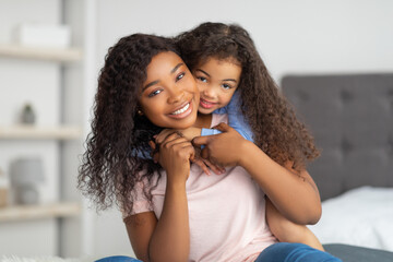 Love between parent and child. Positive little black girl hugging her mom on bed at home