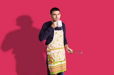 Thoughtful young man wearing kitchen apron looking to camera while holding white blank mock up cup of tea in kitchen and a cooking utensil tools. Coffee break. Cooking pastry for family, dessert