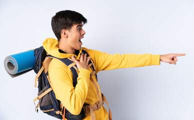 Young mountaineer man with a big backpack over isolated blue background pointing finger to the side