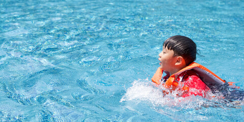 Asian child playing in the swimming pool. Wearing orange life jacket.