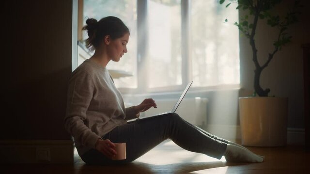 Young Woman Using Laptop at Home for Remote Work. Beautiful Girl Sitting on the Floor Uses Internet, Drinks Morning Cup of Coffee. Sunny Day Ideal for Good for Productive Distant Work