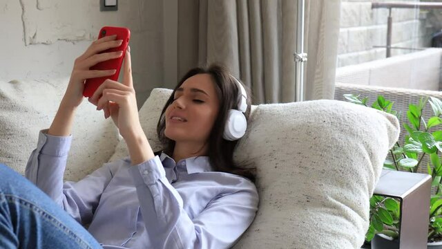 Young attractive smiling caucasian woman using smartphone sitting on sofa at home, singing song, enjoying free time. Girl student studying design apps on digital touchpad.