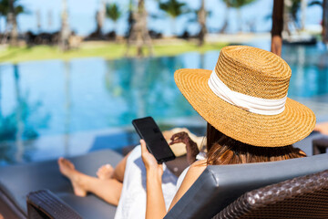 Young woman traveler relaxing and using a mobile phone by a hotel pool while traveling for summer...