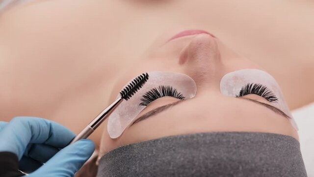 Eyelash extension procedure. Professional beauty master combing female lashes with special mascara brush. Fake eyelashes. Eyelashes extensions close up. Makeup artist and client in beauty salon.