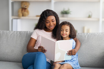 Family stay home pastimes. African American mother and her little daughter reading book together in...