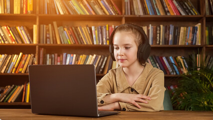 Lady junior student in black headphones listens to teacher and answers sitting at wooden table with grey laptop against racks of books, sunlight