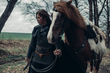 Woman in image of medieval warrior stands near horse among forest.