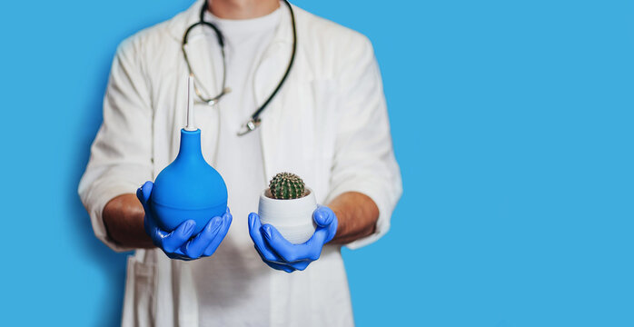 A male doctor holds an enema and a cactus. The concept is the treatment of hemorrhoids, medical care. banner. close-up. A place for text. isolate. medicine. Doctor in a white coat on a blue background