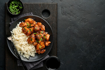 Spicy teriyaki chicken fillet pieces with rice, green onions and black sesame seeds on black plate on a dark slate, stone or concrete background. Top view with copy space.