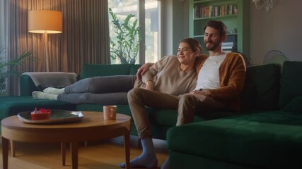 Couple Watches TV together while Sitting on a Couch in the Living Room. Girlfriend and Boyfriend embrace, cuddle, talk, smile and watch Television Streaming Services. Home with Cozy Stylish Interior. - Powered by Adobe