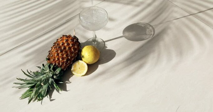 Tropical still life with pineapple and lemon, with shadows from a palm tree and a glass of sparkling drink in which a slice of lemon falls and creates a splash. High quality 4k footage