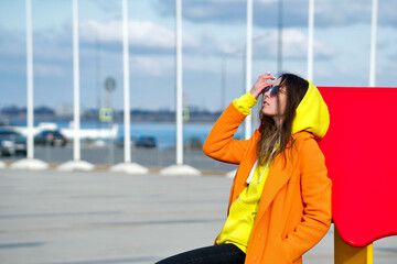 Fototapeta premium Millennial Hipster Girl in lilac sunglasses, orange coat, yellow hoodie posing at urban background. Trendy colorful casual outfit. Street Style. Fashion lookbook concept