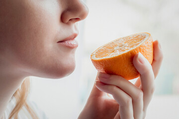 Loss of smell concept. Close up portrait of Caucasian young woman stands near the window and sniffs an orange - obrazy, fototapety, plakaty