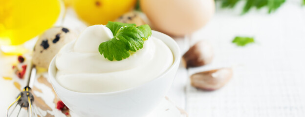 Traditional mayonnaise sauce in white ceramic bowl and ingredients for its preparation on white wooden background. Selective focus