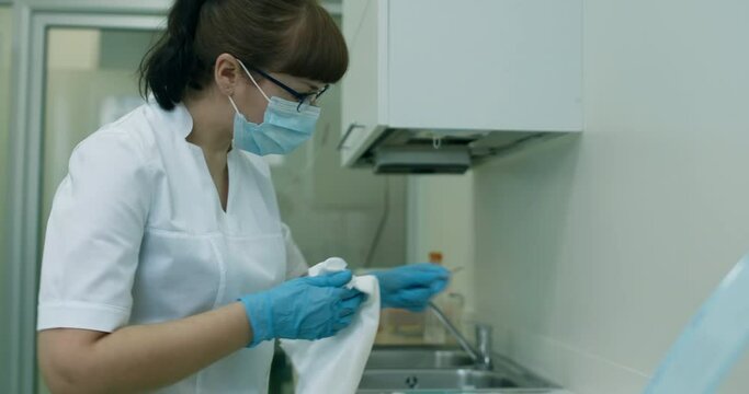A female medical worker in a disposable mask and gloves is wiping a dental tool by using a towel.
