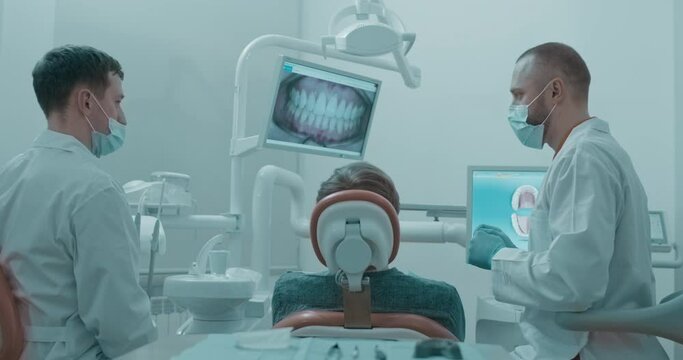 A male dentist and a male assistant are sitting between a male patient that sits in a dental chair and going to the lay position while looking on a screen in front of him with a photo of human teeth
