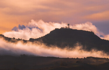 Obraz premium Rocca di Radicofani main tower fortification covered with morning mist. Sunrise light covering the curly clouds running over meadows and fields making light-shadows playing on Tuscany hills landscape.