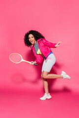 full length of excited african american woman holding tennis racket on pink