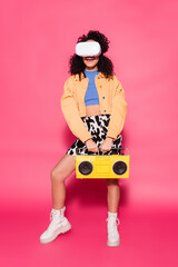 full length of smiling african american woman in vr headset holding boombox on pink