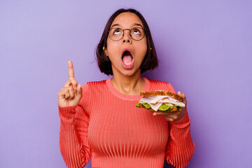 Young mixed race woman holding a sandwich isolated on purple background pointing upside with opened mouth.