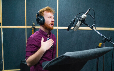 Expressive face of bearded man with red curly hair wear headphones near microphone who makes professional dubbing on a voice recording studio - obrazy, fototapety, plakaty