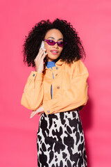 curly african american woman in sunglasses talking on smartphone on pink