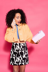 amazed curly african american woman in crop top and jacket talking on smartphone and holding present on pink