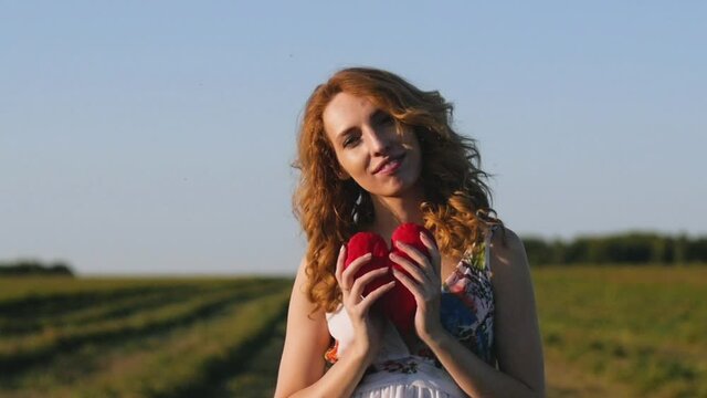 red-haired young woman in a white dress holding a soft heart on the background of the field.