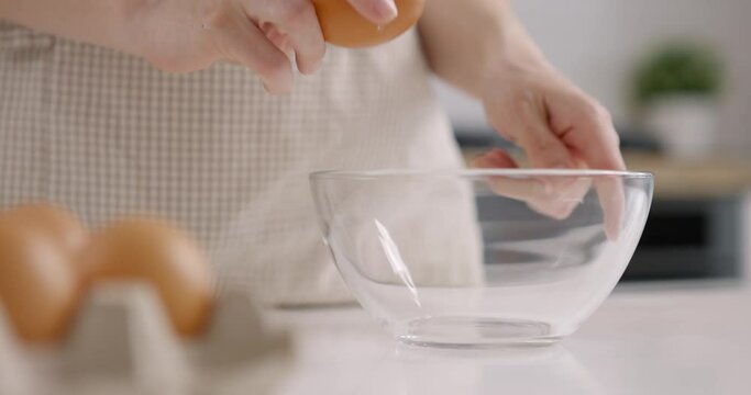 Woman in grey apron takes the egg from the basket and breaks it into glass bowl. Cooking at home kitchen. Slow motion