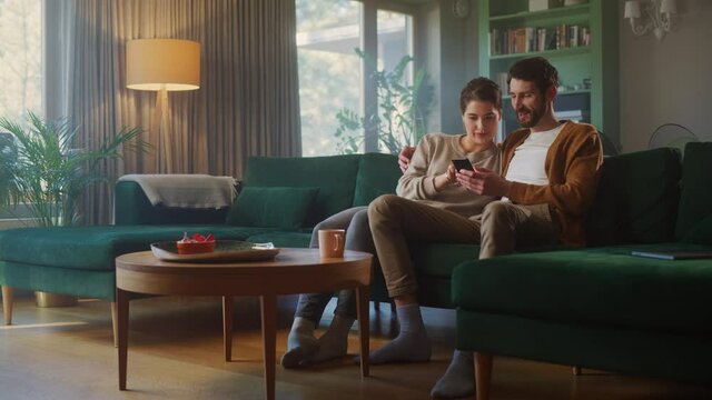 Couple Use Smartphone Device, while Sitting on a Couch in the Cozy Apartment. Boyfriend and Girlfriend Talk, do e-Shopping on Internet, Watching Funny Videos, Use Social Media, Streaming Service.