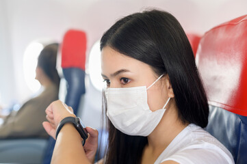 Fototapeta na wymiar Traveling woman wearing protective mask onboard in the aircraft using smart watch, travel under Covid-19 pandemic, safety travels, social distancing protocol, New normal travel concept