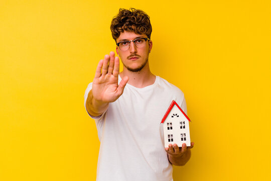 Young caucasian man holding a model house isolated on yellow background standing with outstretched hand showing stop sign, preventing you.