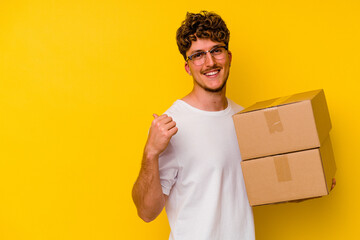 Young caucasian man holding a cardboard box isolated on yellow background points with thumb finger...