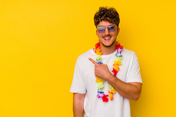 Young caucasian man wearing a Hawaiian party stuff isolated on yellow background smiling and pointing aside, showing something at blank space. - obrazy, fototapety, plakaty