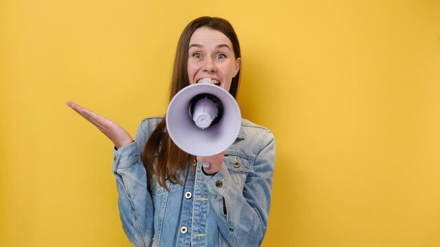 Shocked crazy european young female 20s screaming in megaphone spreading hands, dressed in blue denim jacket, posing isolated over yellow studio background. People sincere emotions lifestyle concept