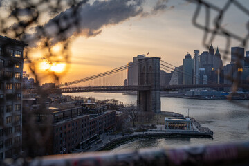 Die Brooklyn Bridge mit der Skyline von New York City im Hintergrund, fotografiert bei Sonnenuntergang von durch einen Zaun auf der Manhattan Bridge