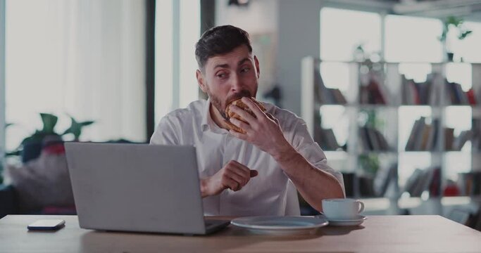 Caucasian excited millennial adut watching fun videocontent on laptop internet social media, eating croissant and drinking coffee in the morning.