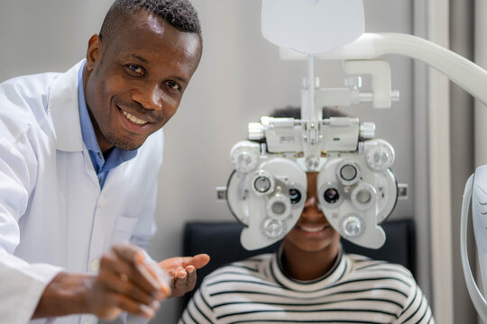 Optician Doing Optometry Eye Exam For Black African Teen Girl Patient. Male Optometrist With Phoropter While Examining Patient.